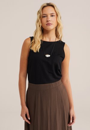 Szőke nő ujjatlan fekete felsőben, barna pliszírozott szoknyában és ezüst medál nyaklánccal, egyszínű bézs háttér előtt.