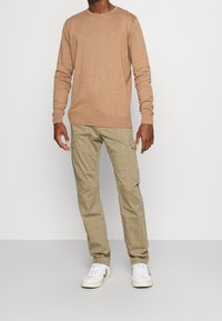 Beżowy wełniany sweter z okrągłym dekoltem, zestawiony z jasnobeżowymi spodniami cargo z wzorem w kratkę, noszony z białymi trampkami.