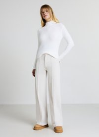 Weißer, eng anliegender Rollkragenpullover kombiniert mit hellbeigen, weit geschnittenen, gerippten Hosen und braunen Schuhen, vor neutralem Hintergrund.