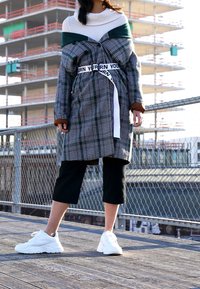 Übergroßes kariertes Mantel mit grünen Akzenten und weißem Gürtel, darüber ein weißes Strickoberteil; kombiniert mit schwarzen, gekürzten Hosen und weißen Sneakers.