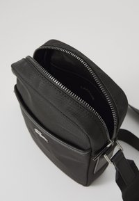 Black nylon crossbody bag with a textured finish, silver zipper closure, and adjustable strap. Features a front pocket and branded logo.