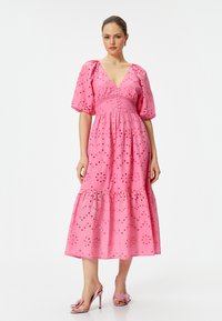 Roze katoenen jurk met korte pofmouwen, V-hals, knoopdetail, borduursels en een gerimpelde zoom. Gecombineerd met bijpassende roze hakken.