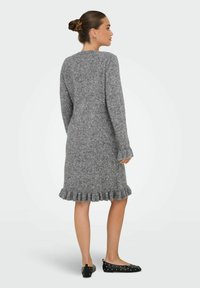 Femme de profil portant une robe tricotée grise jusqu'au genou avec un ourlet et des manches volants, associée à des ballerines noires à clous.