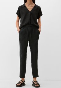 Svart linne kortärmad skjorta med V-ringning, i kombination med svarta byxor med elastisk midja och sidofickor, samt svarta sandaler med remmar.