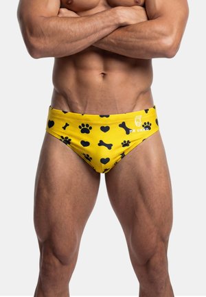 Gespierde torso en benen van een man die gele zwembroek draagt met zwarte pootafdrukken, hartjes en botten, armen over de borst gekruist.