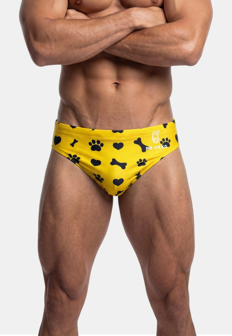 Męski umięśniony tors i nogi w żółtych kąpielówkach z czarnymi nadrukami łap, serc i kości, z rękami skrzyżowanymi na piersi.