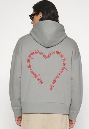 Homme portant un sweat à capuche gris avec un texte rouge en écriture cursive formant un cœur dans le dos qui affiche un message inspiré de la nature.