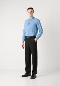 Lichtblauwe blouse met lange mouwen en opstaande kraag, gecombineerd met zwarte broek en gepolijste zwarte schoenen.