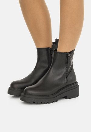 Piernas con botas de tobillo de cuero negro con suelas gruesas y cremalleras laterales, de pie sobre un fondo blanco.