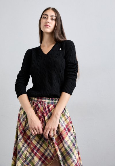 Fiatal nő, hosszú barna hajjal, fekete, kötött V nyakú pulóvert és színes kockás szoknyát viselve, barna táskát tartva egy szürke fal előtt.