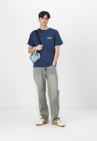 Blå bomulds t-shirt med gul grafik, lys blå crossovertaske, lyse denimjeans og cremede sko med røde striber. Hvid baggrund.