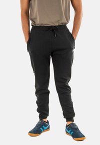 Zwarte sweatpants met een touwtjes taille, zijzakken en een aansluitende boord. Draagt sneakers in marineblauw en turquoise.