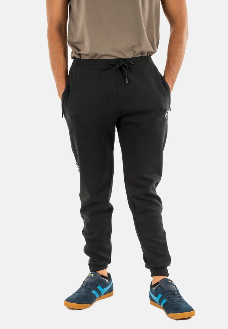 Zwarte sweatpants met een touwtjes taille, zijzakken en een aansluitende boord. Draagt sneakers in marineblauw en turquoise.