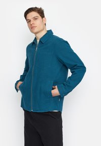 Bermuda groene ritssluiting jasje van textuurstof, met een kraag, twee voorzakken en knoopmanchetten. Geko zen over een lichtgekleurde shirt.