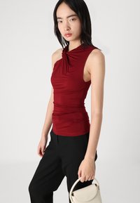 Top sin mangas burdeos con escote retorcido y detalles fruncidos, combinado con pantalones negros y un pequeño bolso blanco.