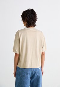 T-shirt beige à manches courtes avec une coupe décontractée, en tissu doux. Stylé avec un col rond et légèrement court au niveau de la taille, associé à un jean bleu.