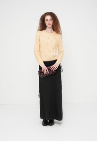 Pull jaune pâle en tricot avec broderies florales, associé à une longue jupe noire. Tenant une pochette bordeaux foncé. Bottines noires.