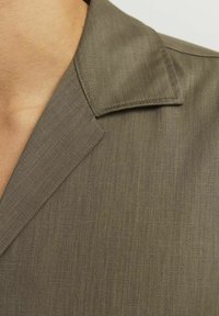 Chaki marškiniai su plačiu apykakle, pasižymintys lygiu tekstūravimu ir subtiliu įstrižiniu raštu. Artimas apykaklės ir peties vaizdas.