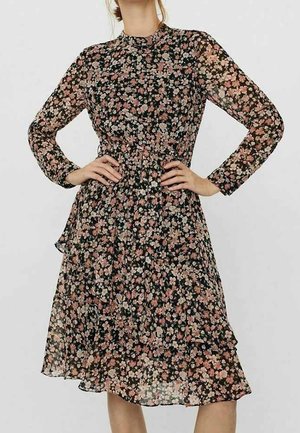 Nainen pukeutuneena polvipituiseen mustaan mekkoon, jossa on pitkät läpikuultavat hihat ja vaaleanpunainen kukkakuosi, kädet lanteilla yksinkertaisen taustan edessä.