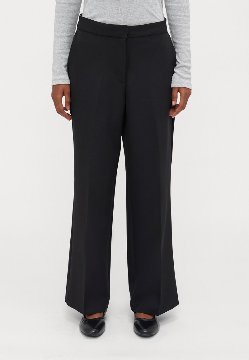 Personne portant un pantalon noir à jambes larges, un haut gris à manches longues et des chaussures noires, debout devant un fond uni.