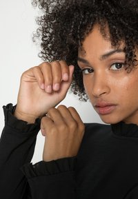 Zwarte blouse met lange mouwen en ruches aan de manchetten, met een gladde textuur. De handen zijn nabij het gezicht gepositioneerd en tonen natuurlijke huidskleuren.