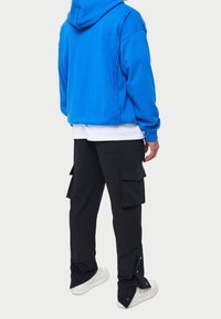 Niebieska bluza z kapturem o luźnym kroju, zestawiona z czarnymi spodniami cargos z wieloma kieszeniami i zapięciami na zatrzask, noszona z białymi trampkami.