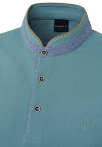 Polo-Shirt aus türkisfarbenem Stoff mit einem kragen ähnlichen Denim, drei Knöpfen und einem kleinen gestickten Logo am Saum. Glatte Textur.