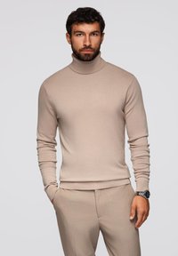 Maglione beige a collo alto realizzato in morbido tessuto a maglia, con maniche lunghe e un design aderente, abbinato a pantaloni sartoriali e un orologio.