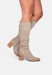 Beige Wildleder-Stiefel in Knielänge mit tailliertem Design, abgerundeter Spitze und strukturiertem Holzabsatz. Mit einem seitlichen Reißverschluss für einfaches An- und Ausziehen.
