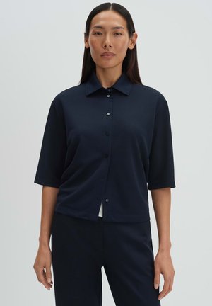 Frau mit langen dunklen Haaren, die ein dunkelblaues, kurzärmeliges Hemd mit Knöpfen und passende Hosen trägt, steht vor einem schlichten grauen Hintergrund.