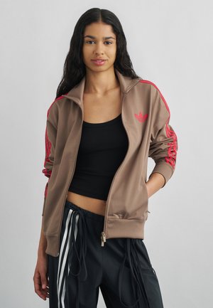 Junge Frau mit langen dunklen Haaren, die eine braune Trainingsjacke mit roten Streifen, ein schwarzes Crop-Top und schwarze Hosen mit weißen Streifen trägt und vor einem grauen Hintergrund posiert.