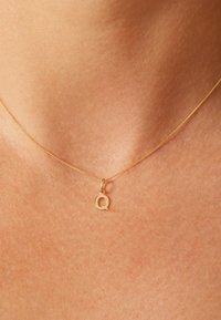 Gold necklace with a fine chain, featuring a small "Q" pendant. The pendant is simple and unadorned, resting on bare neck skin.