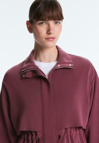 Young woman with straight brown hair in a bun wearing a maroon zip-up jacket over a white top, looking slightly to the side.