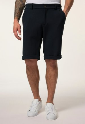 Uomo che indossa pantaloncini neri con orlo arrotolato e sneakers bianche, in piedi con una mano in tasca e tatuaggio visibile sul braccio sinistro.