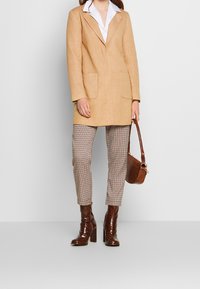 Abrigo beige con solapa, dos bolsillos delanteros; llevado sobre pantalones marrones de cuadros. Combinado con botas de tacón marrones brillantes y un bolso.