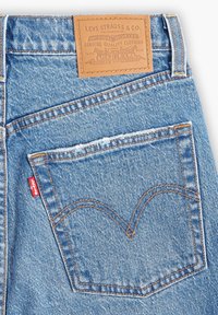 Jeans de mezclilla azul con un parche de cuero marrón con logo en relieve, costuras naranjas y una etiqueta roja en el bolsillo izquierdo.