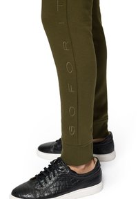 Oliwkowe dresy z napisem "GO FOR IT" w jasnobrązowym kolorze po boku, zestawione z czarnymi, teksturowanymi butami sportowymi z białymi podeszwami.