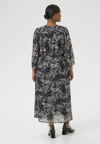 Robe longue et fluide en tissu transparent avec un motif abstrait bleu marine et blanc, dotée de longues manches et d'une taille élastique, accompagnée de talons noirs.