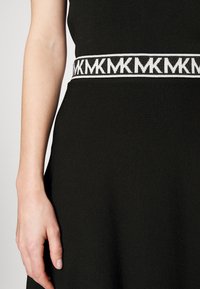 Robe noire avec une sangle tissée à motif présentant un logo "MK" répété en blanc. Tissu lisse avec une coupe ajustée montrée sur un bras.