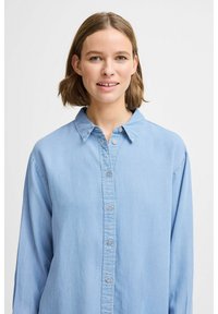 Camicia button-up azzurro chiaro in morbido tessuto, con colletto classico e maniche lunghe, dotata di una chiusura a bottoni frontale completa e un design minimalista.
