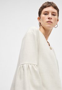 Ανοιχτό μπεζ, υφασμάτινη μπλούζα με υφή και V λαιμόκοψη, φουσκωτά μανίκια και μία χρυσή λεπτομέρεια με κουμπί.