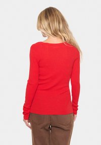 Red ribbed long-sleeve sweater with a rounded neckline. Fitted design with a smooth texture, showcased from the back. Paired with brown pants.