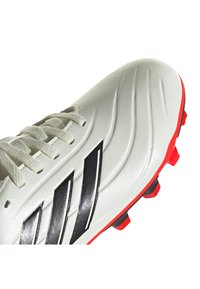 Botín de fútbol blanco con rayas negras, parte superior de cuero texturizado y suela exterior roja vibrante. Presenta cordones tradicionales y acentos de diseño angulares.