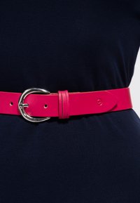 Leuchtend pinker Ledergürtel mit glänzender Oberfläche, ausgestattet mit einer silbernen ovalen Schnalle und zwei kleinen Löchern zur Verstellung. Getragen über einem dunklen Stoff.