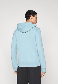 Ανοιχτό μπλε φούτερ με κουκούλα και κορδόνι, με ριμπ μανσέτες και τελείωμα. Λείο ύφασμα με άνετη εφαρμογή, φορεμένο με μαύρο παντελόνι.