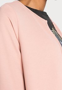 Person som bär en texturerad ljusrosa kofta över en mörk grafisk T-shirt, fångad från axeln till mitten av bröstet.