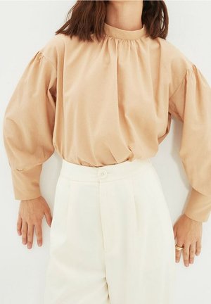 Frau trägt eine beige Bluse mit Puffärmeln, die in hoch taillierte weiße Hosen gesteckt ist, steht mit entspannt an den Seiten hängenden Händen.