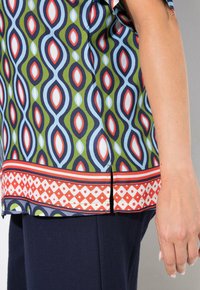 Kurzärmlige Bluse mit Muster in Grün, Blau, Rot und Weiß, mit geometrischem Design und dekorativem Saum, ausgestattet mit Seitenschlitzen für eine bessere Bewegungsfreiheit.