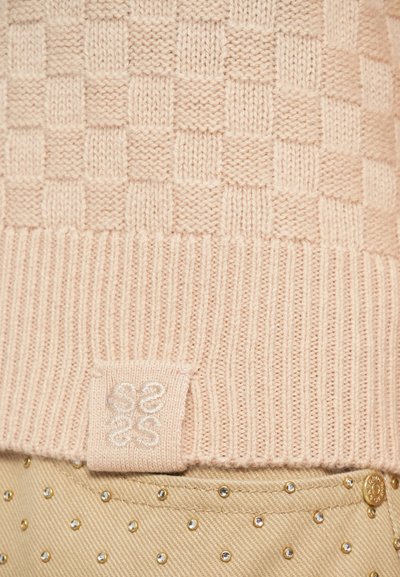 Jasnoróżowy sweter dziany z wzorem w kratkę, z prążkowanym wykończeniem i małą metką z logo. Połączony z beżowymi spodniami ozdobionymi małymi złotymi ćwiekami.