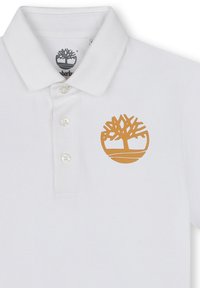 Polo blanc en coton avec un col structuré, arborant un logo d'arbre doré sur la poitrine gauche et une patte de boutonnage à trois boutons.